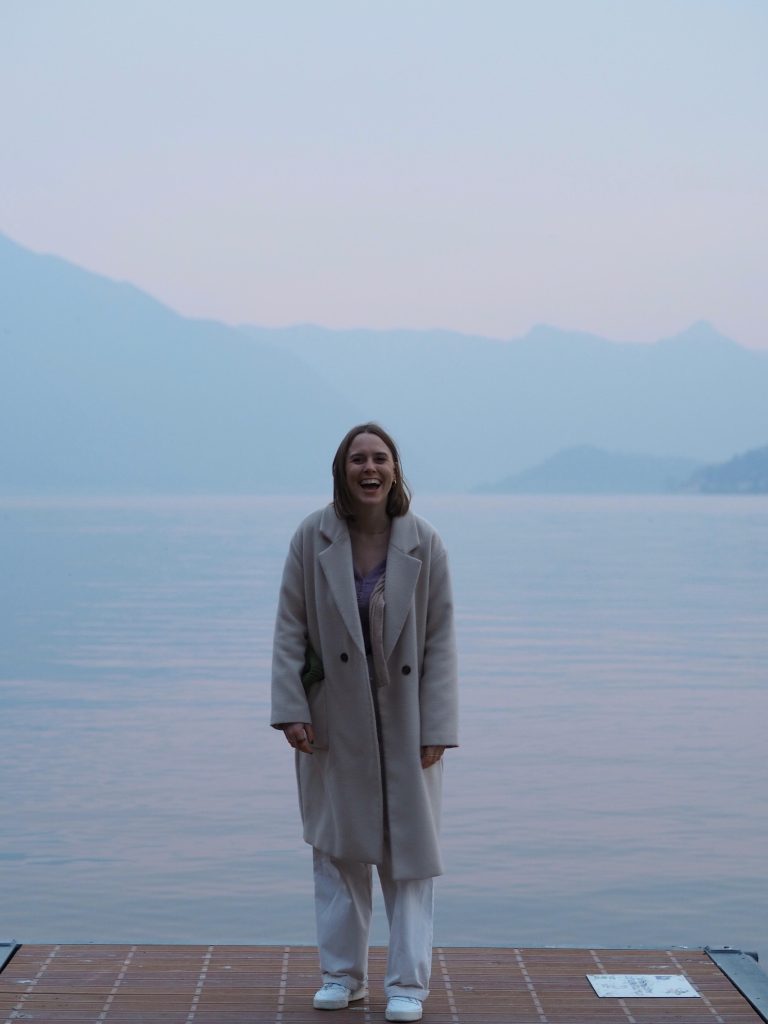 Mujer de pie frente al mar con abrigo claro en día nublado